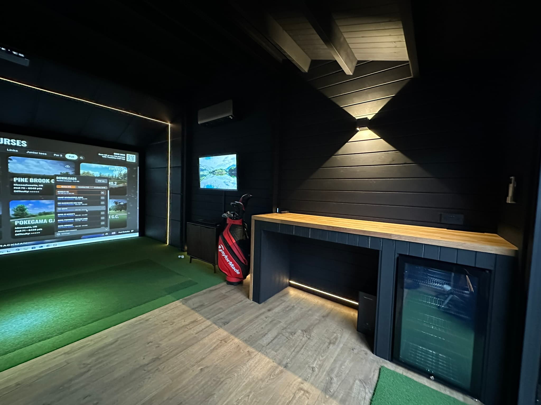 Simulator screen beside a bar counter with a drinks fridge, a golf bag and wall lights