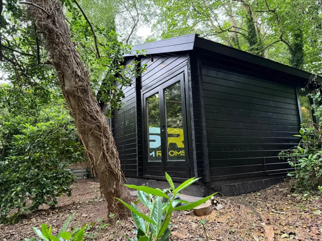 Black-painted log cabin among trees, with the GSR logo seen through its glass doors