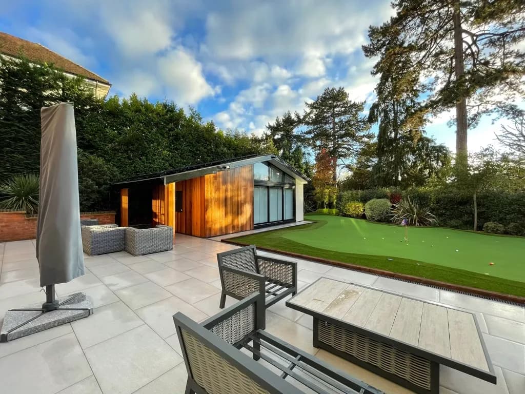 Paved patio with outdoor furniture and a parasol, leading to a clad garden room and a putting green