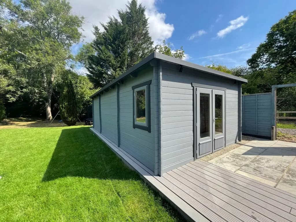 Grey-painted log cabin with double doors and a decked path on a sunny lawn