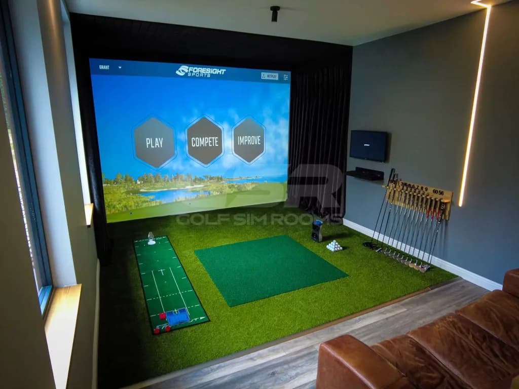 Simulator bay with a course menu on the screen, putting and hitting mats and a rack of clubs, seen past a leather sofa
