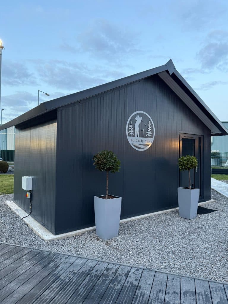 Dark grey garden building with a golfer emblem on the wall and potted trees on gravel at dusk