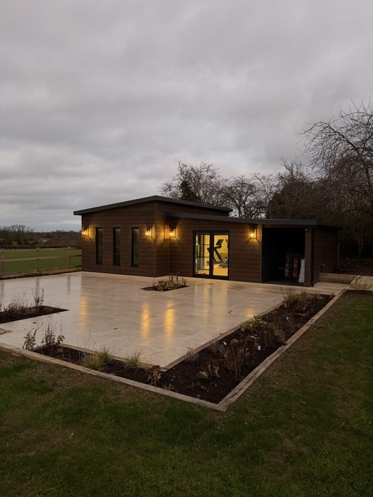 The garden building at dusk seen across the lawn, with planted borders around the patio
