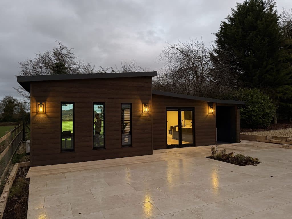 Dark-clad garden room with wall lights and glass doors, behind a stone patio at dusk