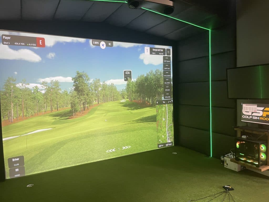 Simulator with green LED edging showing a pine-lined fairway, with a gaming PC on a wall shelf