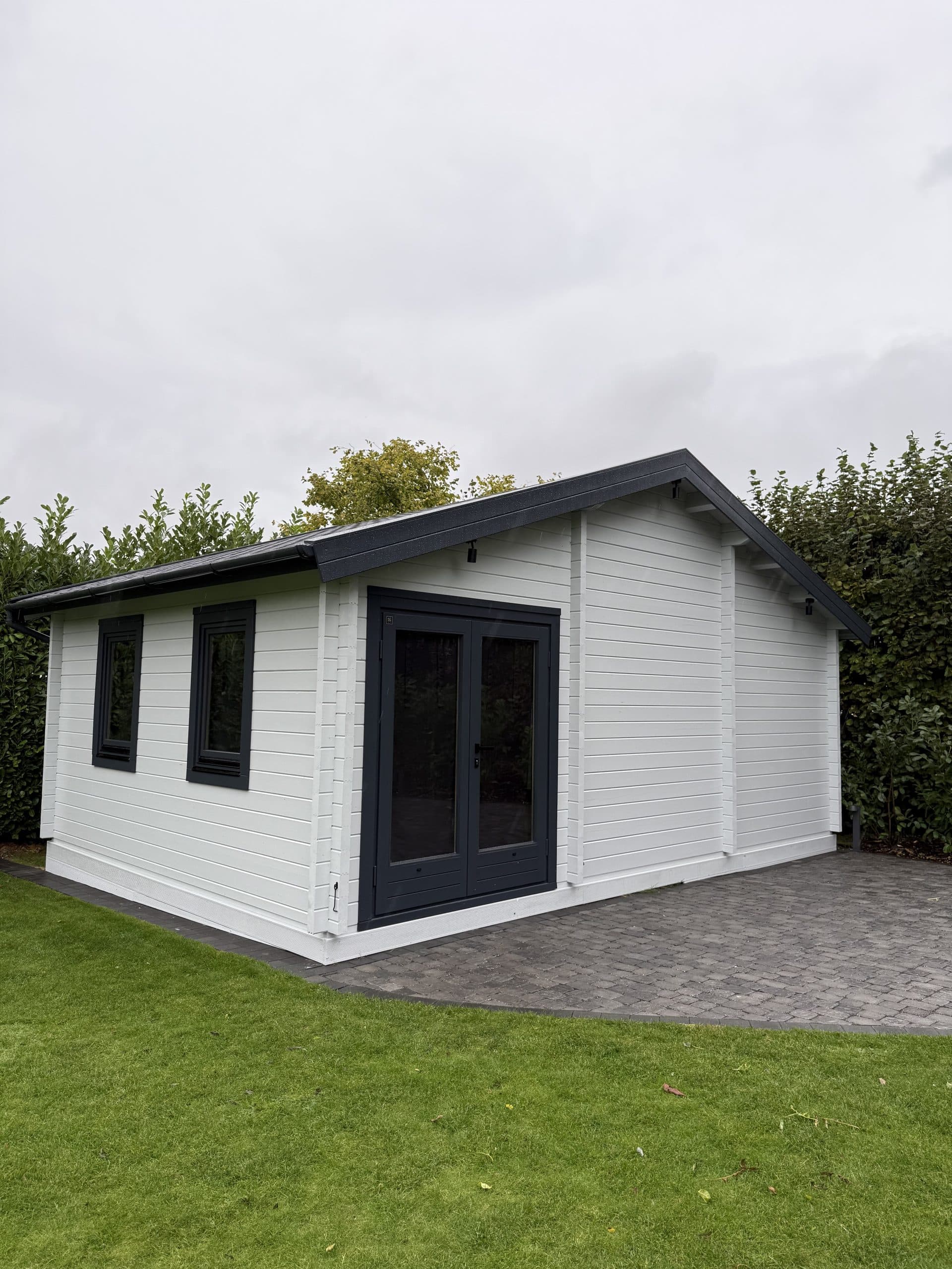 Side view of the grey log cabin, bordered by a hedge