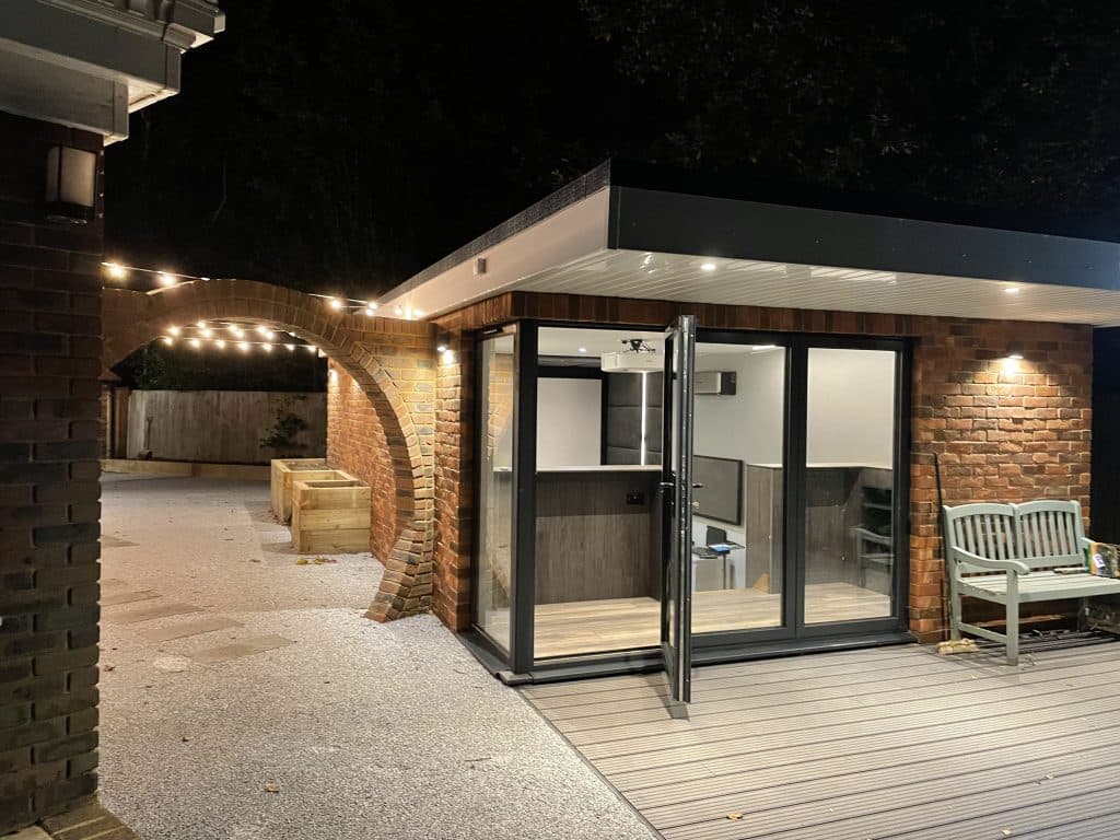 Garden room with open glass doors at night, beside a brick arch strung with festoon lights