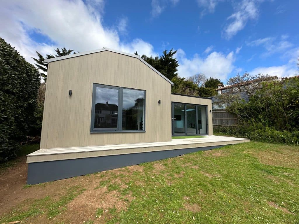 Light-clad garden room with a pitched roof and a flat-roofed wing, on decking in a garden