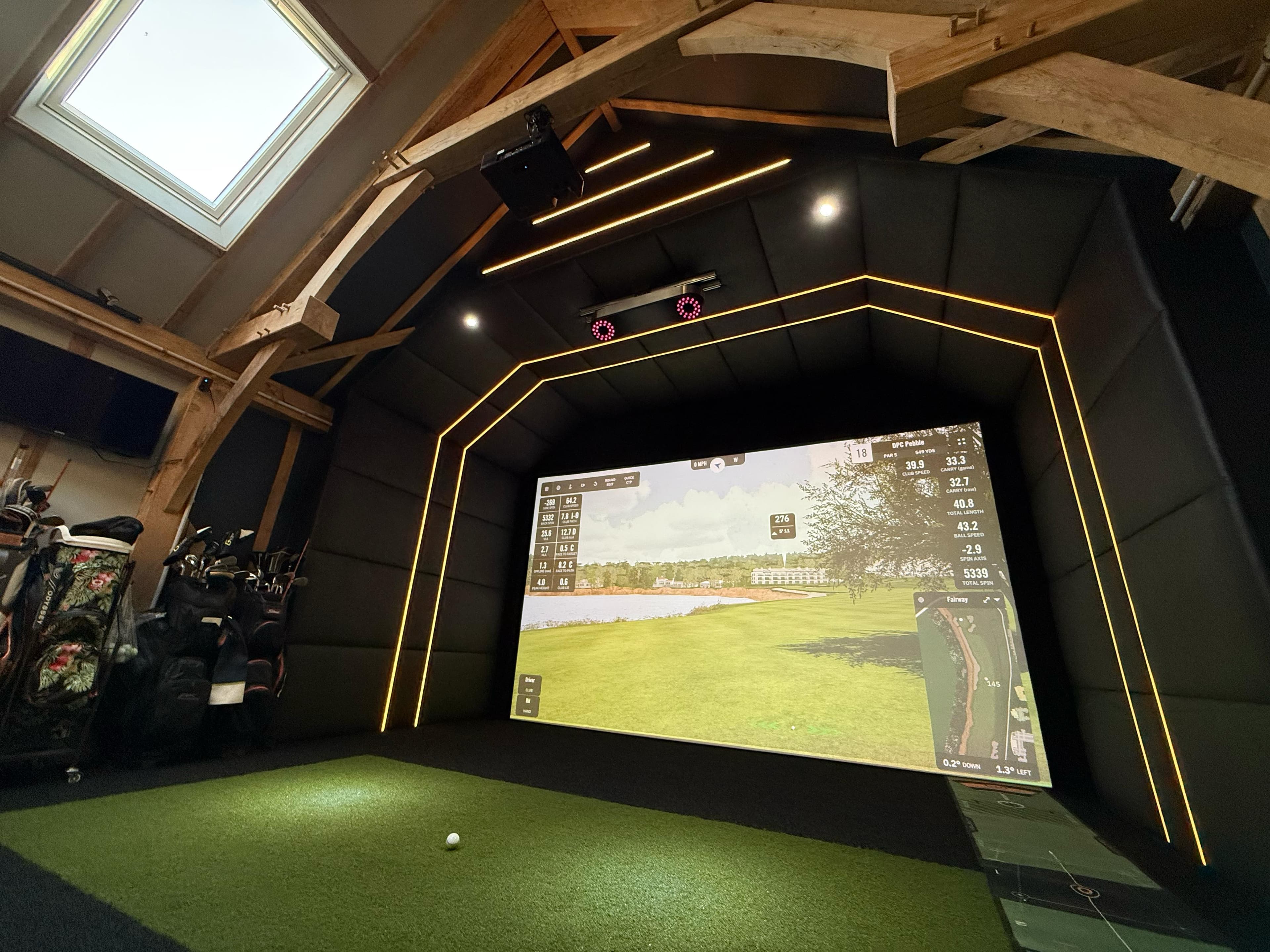 Simulator bay under a skylight, amber LED lines around the screen and golf bags to the left